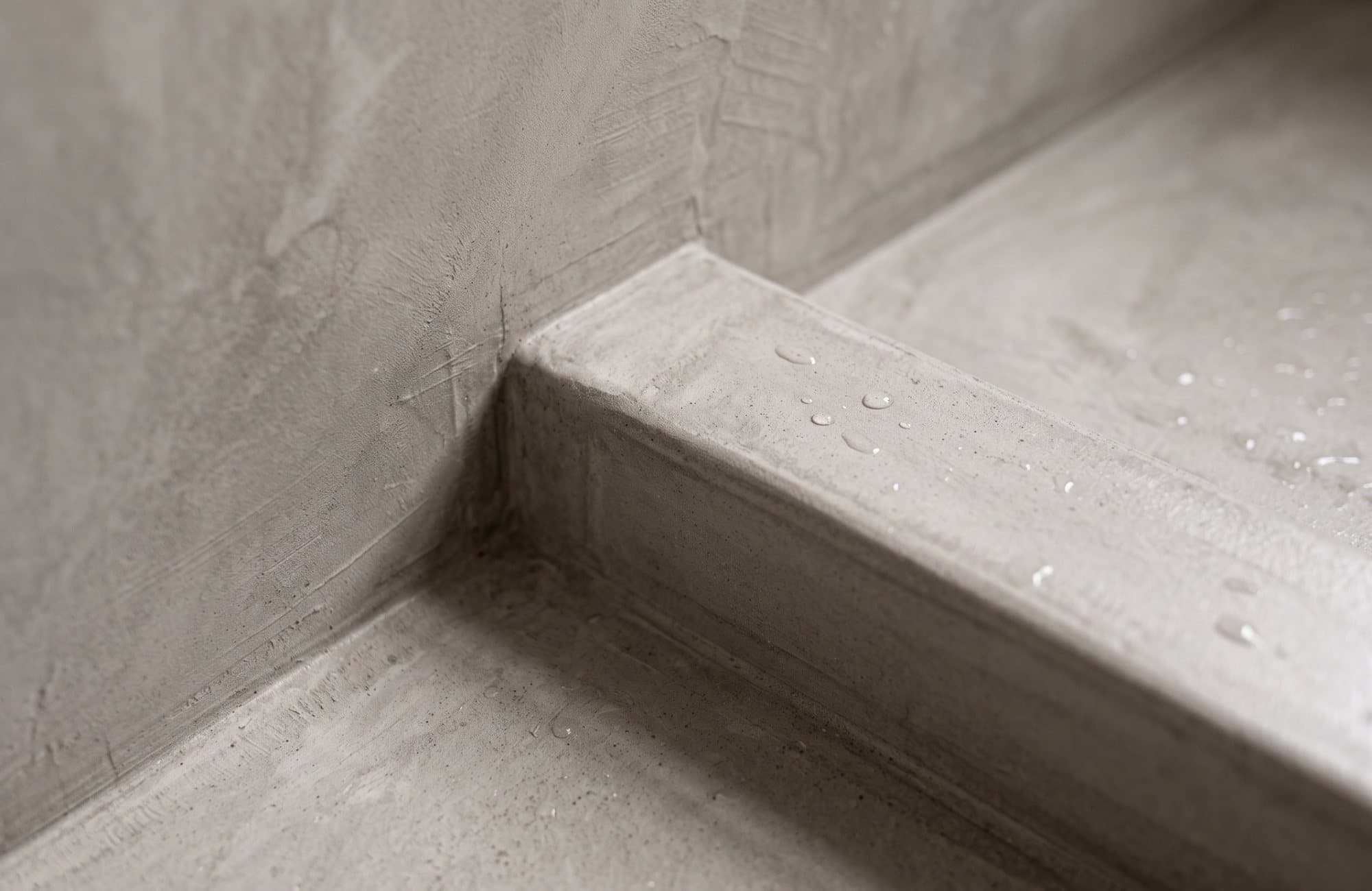 Close-up of the microcement where the shower wall meets the curb, with water beading on the seal
